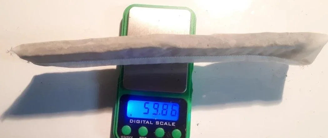 A long pre-rolled paper cone resting on a green digital scale displaying 59.86 grams.